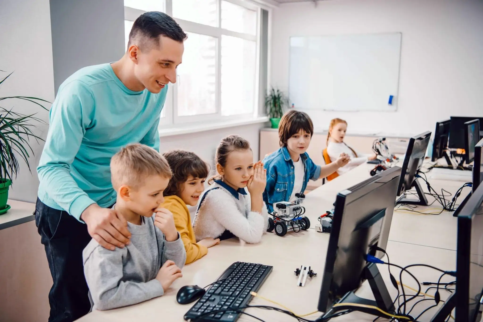 הגנת פרטיות תלמידים בלמידה מרחוק – אתגר הקורונה. התמונה מציגה כיתת לימוד, שבה מורה מדריך ילדים צעירים בלימוד טכנולוגיה ורובוטיקה. הילדים נראים מתעניינים וקשובים למסכים ולציוד הרובוטי שעל השולחן.